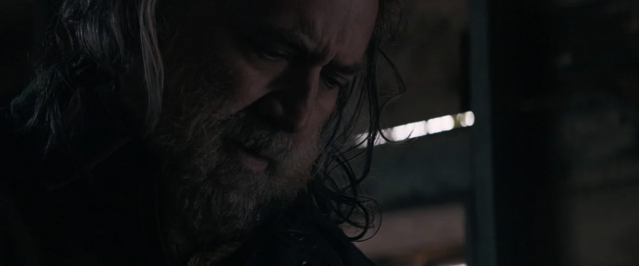 Pig (2021) movie still: closeup — A dimly lit interior space features dark industrial surfaces and a sliver of light. An older man with long hair…