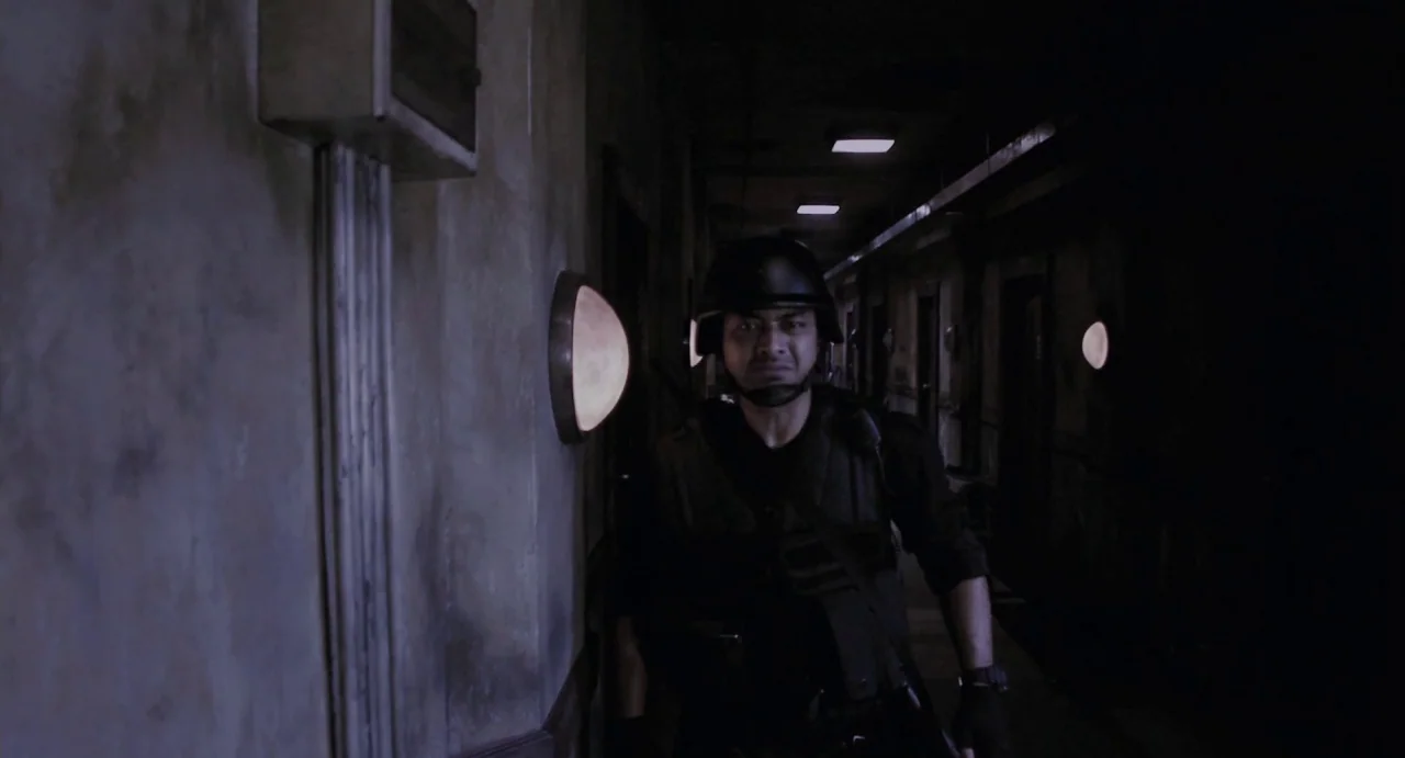 The Raid (2012) movie still: medium — A dimly lit narrow corridor features textured gray walls with mounted spherical wall lights. A man in tactical…