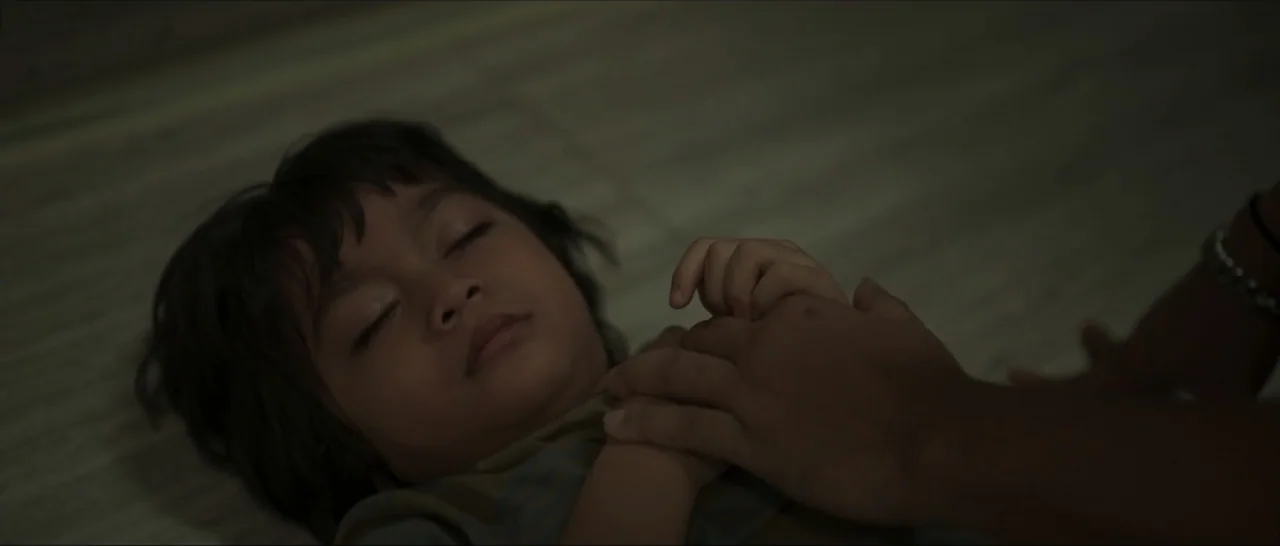 Innocent Vengeance (2023) movie still: closeup — A dimly lit room focuses on a child sleeping on a textured surface. An adult hand is placed gently…