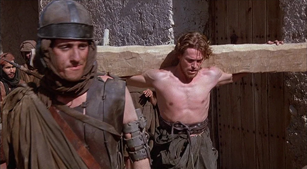 The Last Temptation Of Christ (1988) movie still: medium — A dusty ancient street features textured stone walls and a rustic wooden doorway. A man…