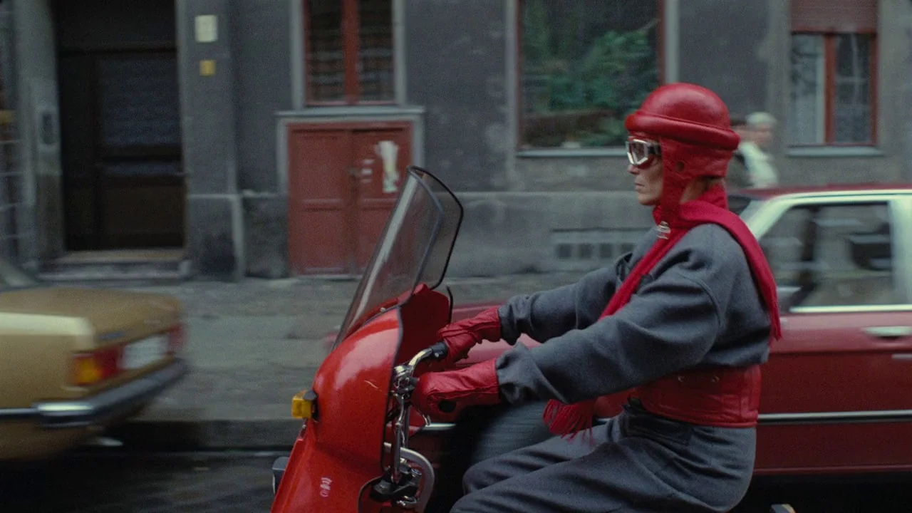 Dorian Gray in the Mirror of the Yellow Press (1984) movie still: medium — A person rides a bright red scooter past a blurred gray urban building…