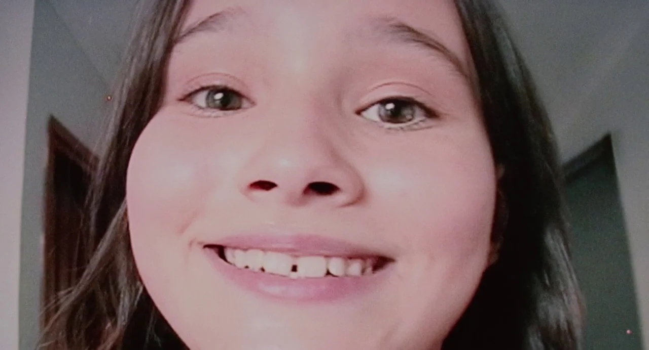 Aftersun (2022) movie still: closeup — A close-up of a young girl inside a dimly lit room. She looks directly into the camera while smiling.