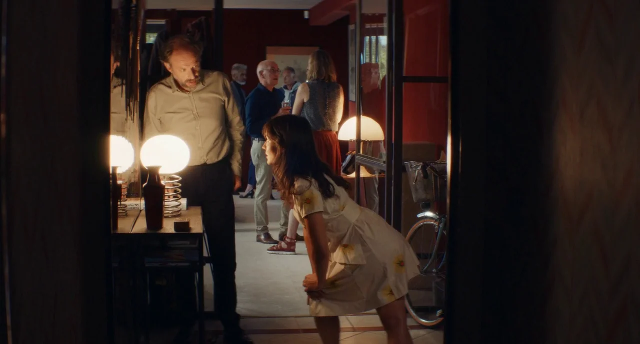 Anaïs in Love (2021) movie still: medium — A dimly lit interior hallway with dark red walls and multiple guests gathered behind. A man stands beside…