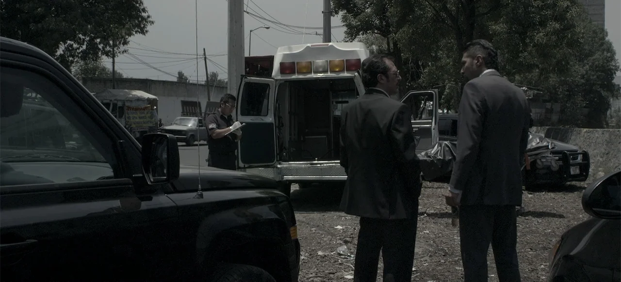 About the Deaths (2016) movie still: fullbody — An outdoor dirt lot contains parked vehicles including a white ambulance. Two men in formal business…