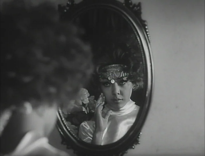 Funeral Parade of Roses (1969) movie still: closeup — An oval-framed wall mirror displays a woman in a white satin top. The woman gazes at her own…