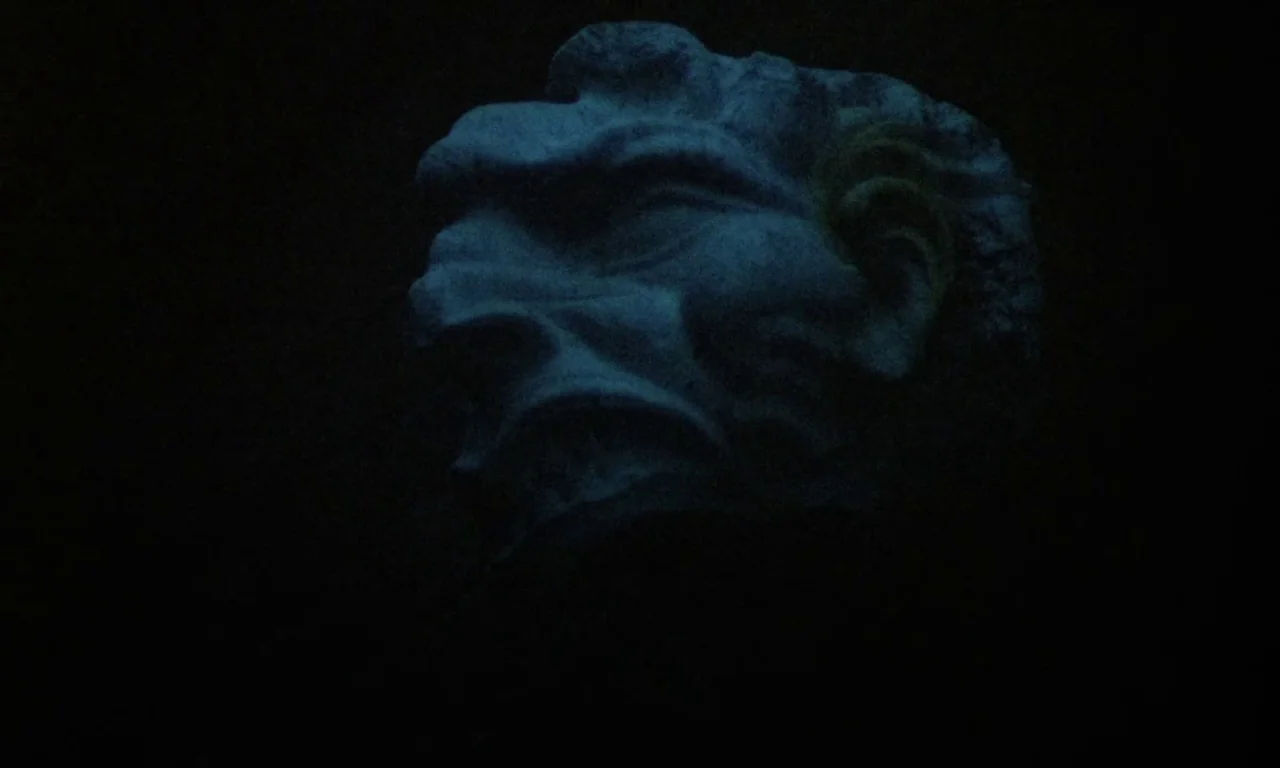 Paganini Horror (1989) movie still: closeup — A stone sculpture of a face is partially visible against a pitch black background. The weathered…