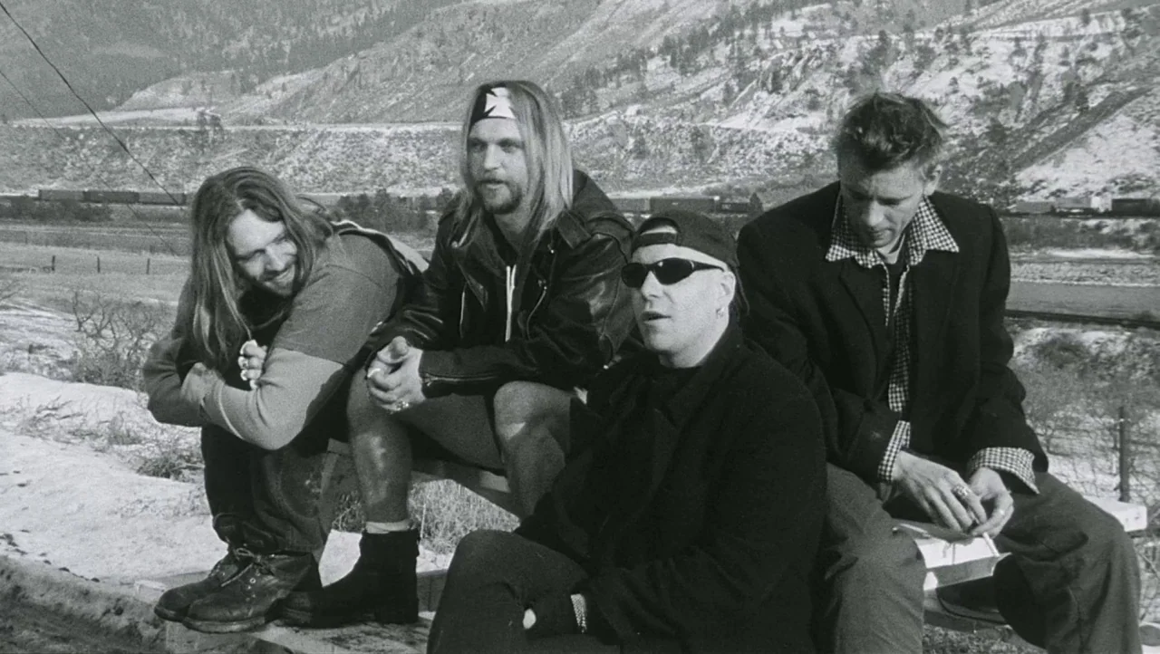 Hard Core Logo movie still: medium — A rural landscape features a snowy hillside and a train track in the background. Four men are seated on a…