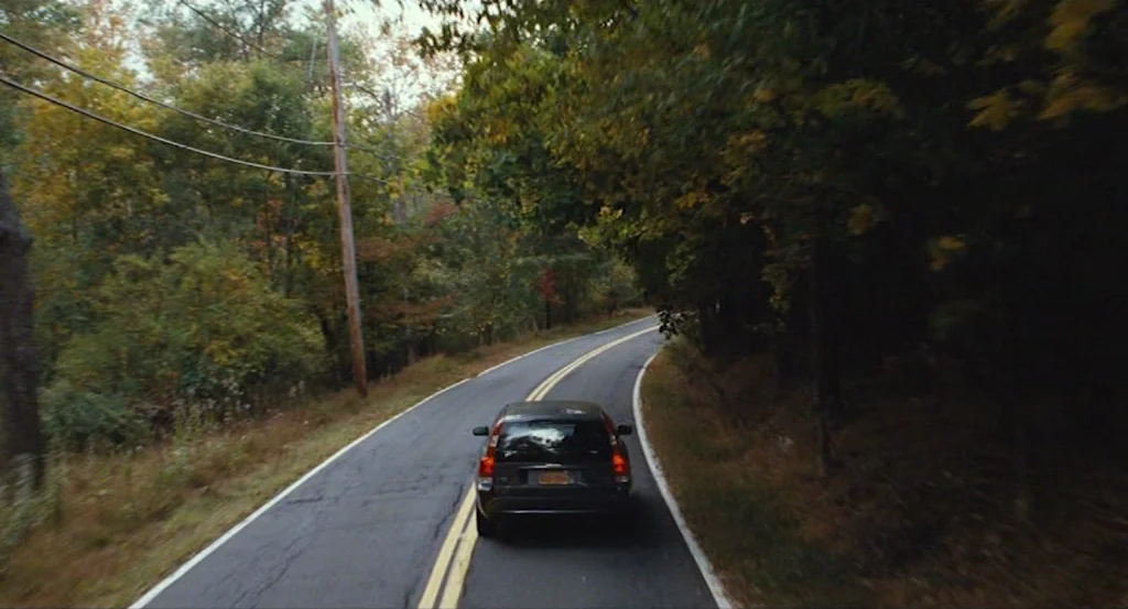 Louder Than Bombs (2015) movie still: wide — A rural asphalt road curves through a dense forest with autumn-tinged deciduous trees. A dark station…