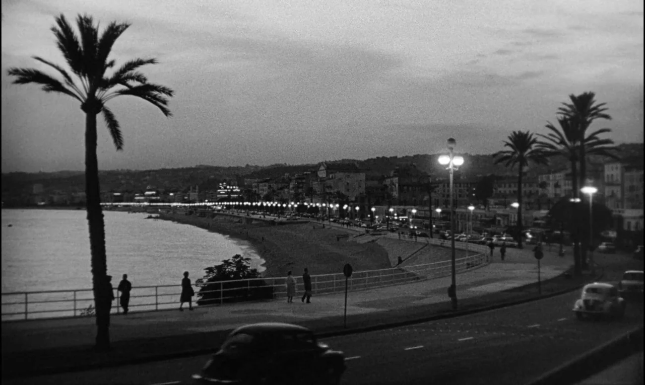 Bay of Angels (1963) movie still: establishing — A curved seaside promenade features tall palm trees and bright streetlamps overlooking the water.…