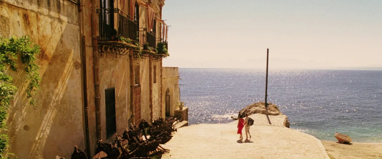 Ocean’s Twelve (2004) movie still: wide — An weathered stone building with iron balconies stands beside a sunlit stone quay. A couple walks hand in…