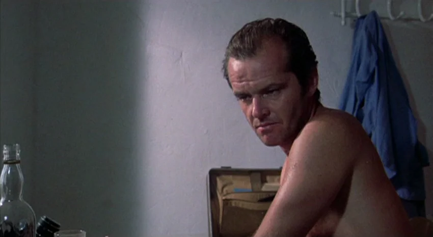 The Passenger (1975) movie still: medium — A bare room features a white wall with a blue shirt hanging on a hook. A shirtless man sits at a table…