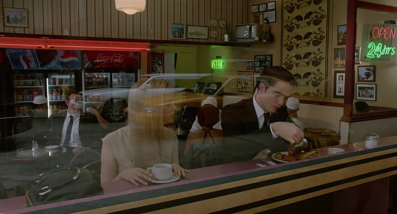 Cosmopolis (2012) movie still: medium — A diner booth with patterned wallpaper and a reflection of a street scene outside. A man and a woman sit at…