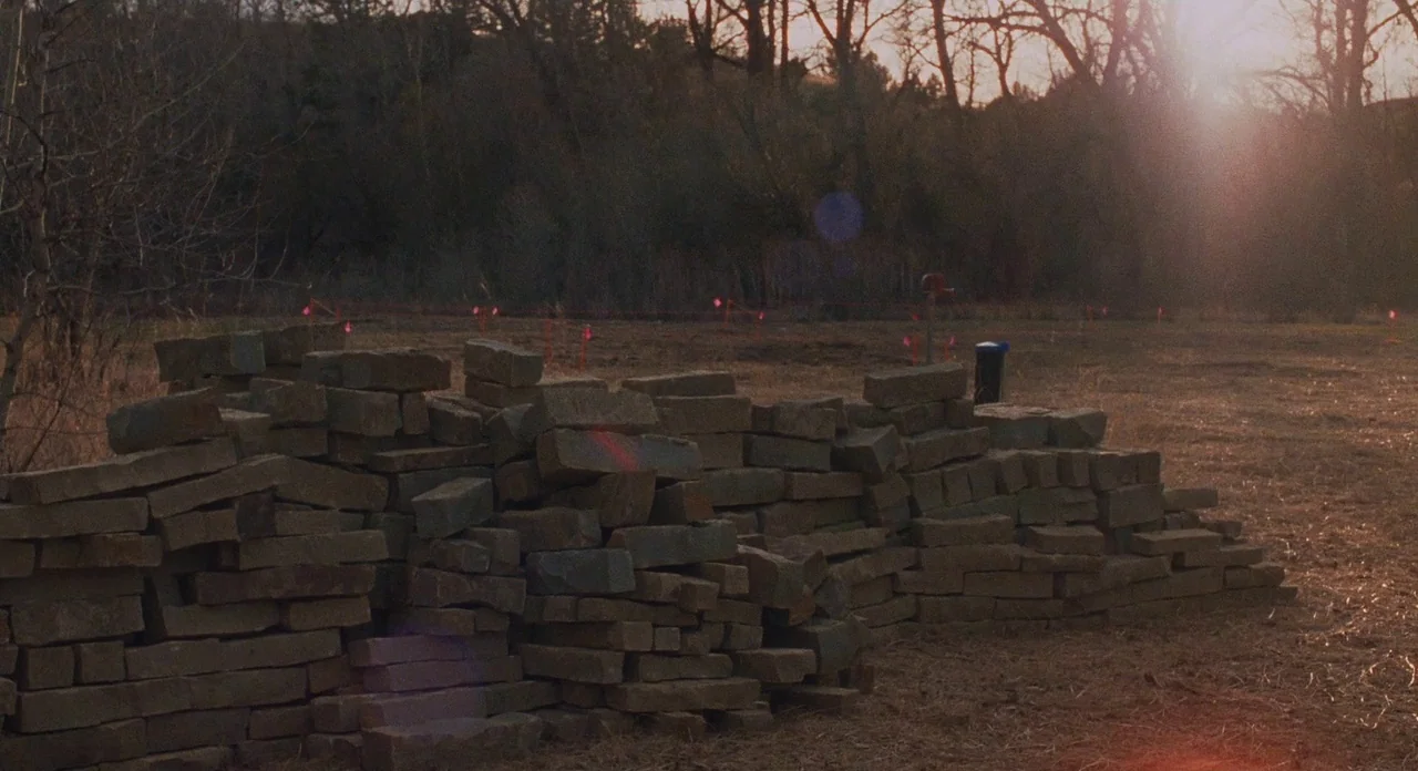 Certain Women (2016) movie still: wide — A pile of rectangular stone blocks sits on a dirt lot at sunset. Bare trees and scattered orange marker…