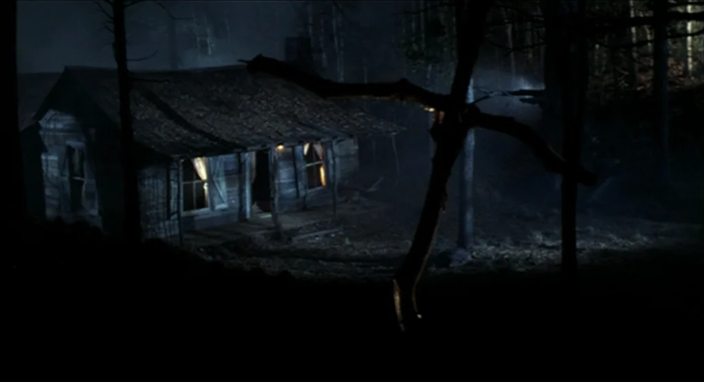 Evil Dead II (1987) movie still: establishing — A wooden cabin sits surrounded by dark, dense trees at night. Faint yellow light spills from the…