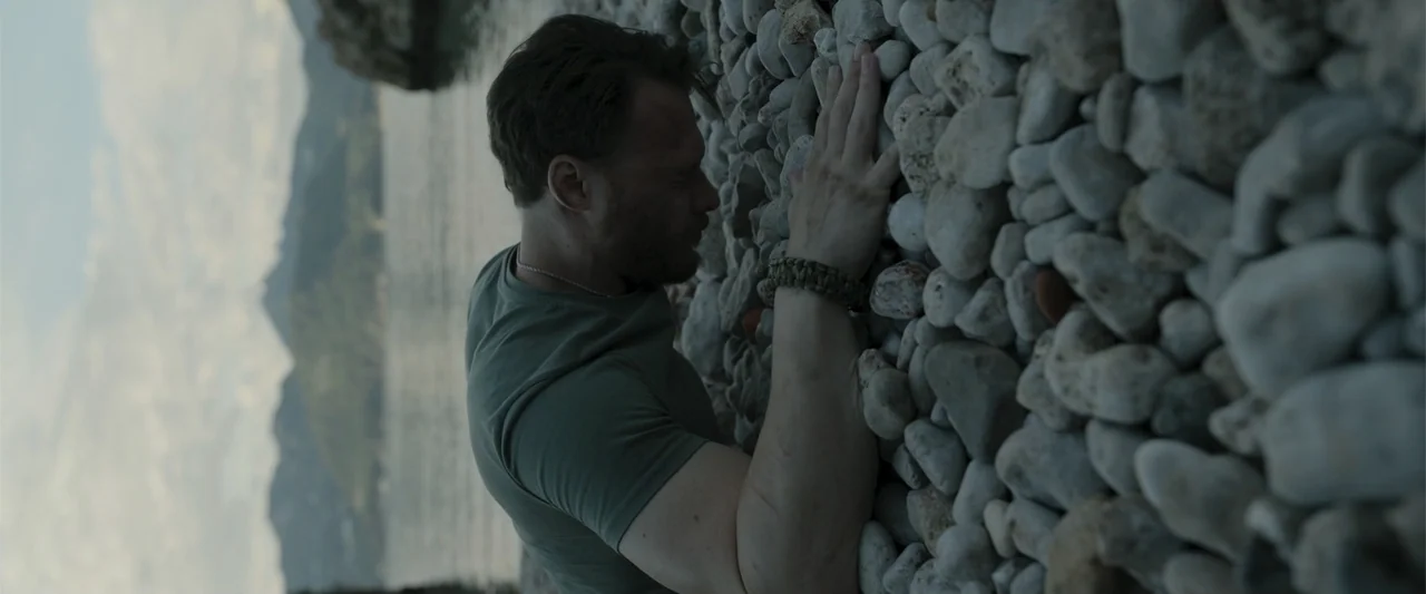 Blue Cave (2024) movie still: medium — A man stands outdoors near a body of water by a large wall of stones. The man in a green t-shirt touches the…