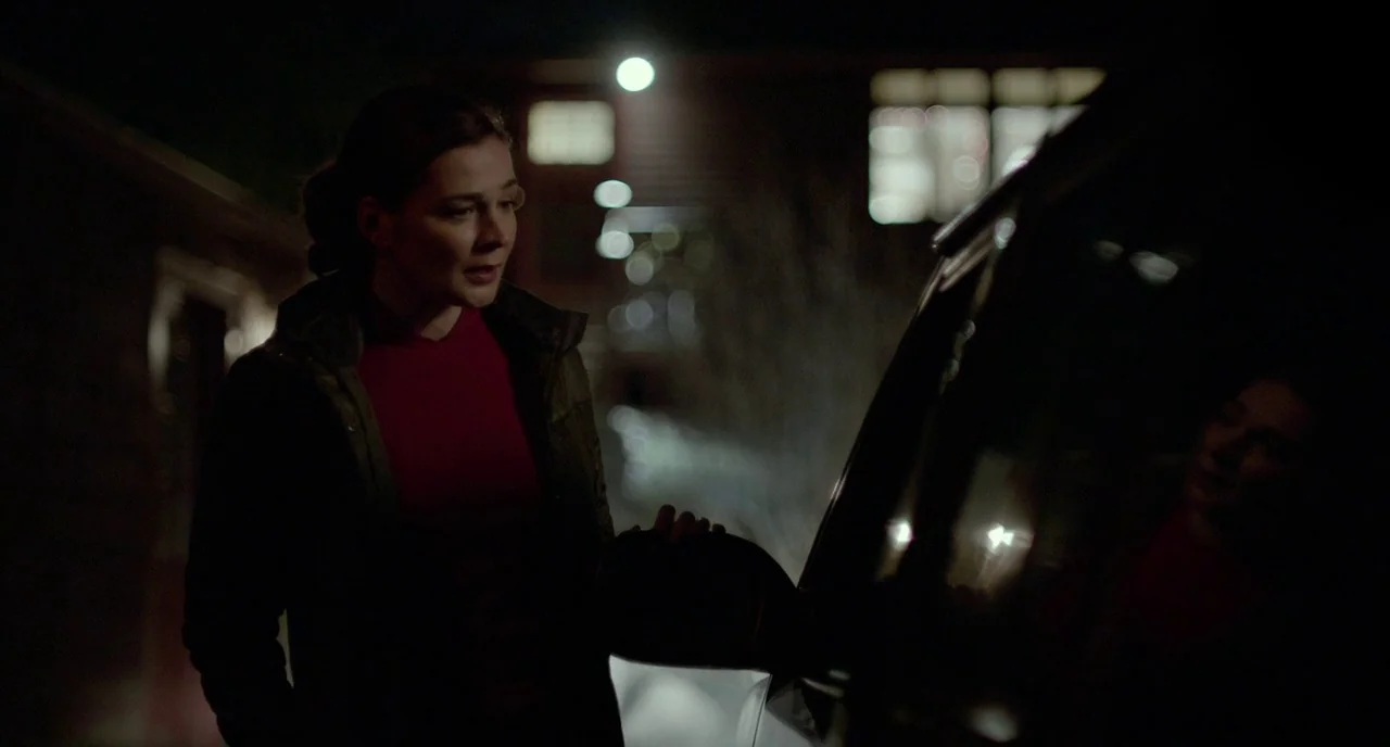 Manchester By The Sea (2016) movie still: medium — A dark outdoor parking lot at night with a blurred building in the background. A woman in a red…