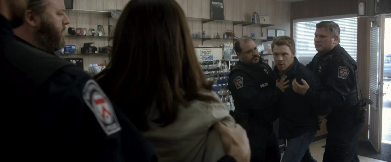Radius (2017) movie still: medium — A cramped store interior features shelves stocked with office supplies and light wood paneling. Two uniformed…