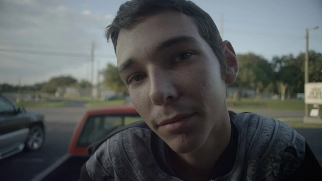 Florida Man (2015) movie still: closeup — A paved parking lot with utility poles and blurred trees under a bright sky. A young man stands close to…