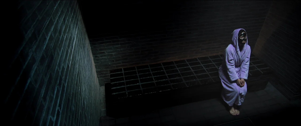 The Feast (2021) movie still: wide — A dark tiled sauna features walls constructed of horizontal rows of dark gray bricks. A woman wearing a…