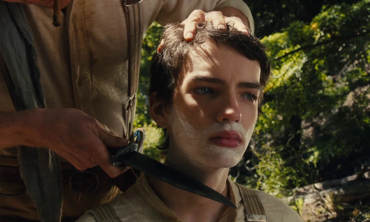 Slow West (2015) movie still: medium — A sunlit woodland clearing provides the setting for a rustic grooming scene. An older man carefully scrapes…