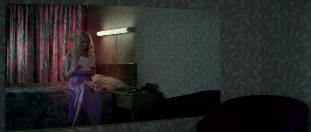 The Neon Demon (2016) movie still: medium — A dimly lit room with patterned wallpaper and a mirror reflecting the bed. A young blonde woman sits on…