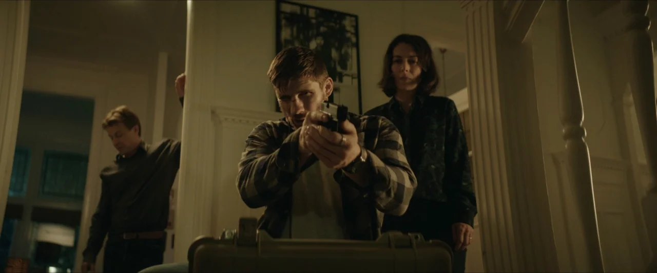 Black Lotus (2023) movie still: medium — A residential hallway at night with white-painted walls and a dark framed picture. One man holds a handgun…