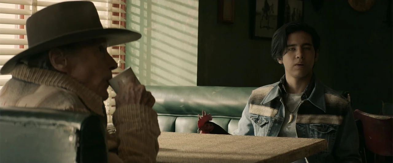 Cry Macho (2021) movie still: medium — An elderly man and a young man sit across from each other in a booth. The booth is green vinyl and a rooster…