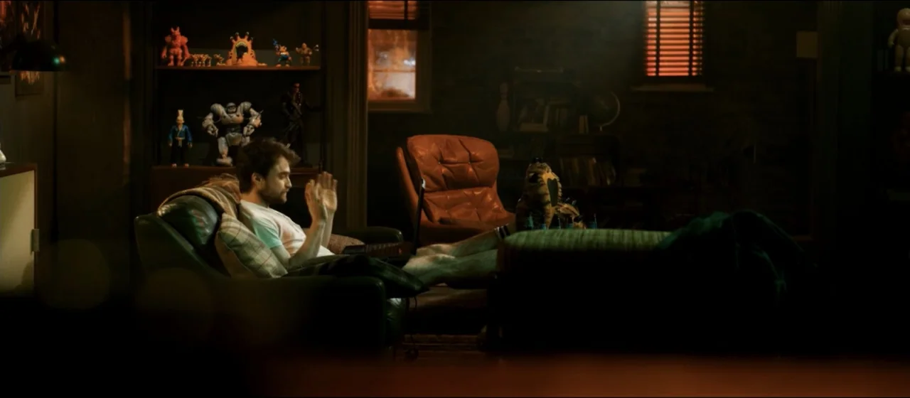 Guns Akimbo (2020) movie still: medium — A dimly lit room contains a leather armchair, a shelf with figurines, and furniture. A young man sits on a…