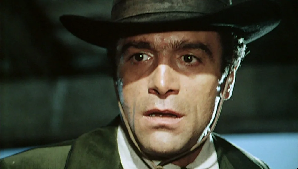 The Great Silence (1968) movie still: closeup — A man in a wide-brimmed hat stands in a dark, dimly lit space. He looks toward the side of the frame…