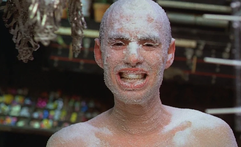 Sweet Movie (1974) movie still: closeup — A close view of a man standing before a cluttered market stall background. The man has his head and upper…