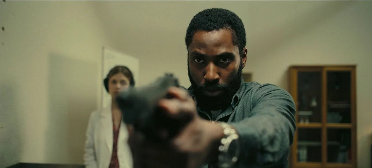 Tenet (2020) movie still: medium — An interior space with plain, neutral-colored walls and a wooden cabinet in the back. A man with a beard holds a…