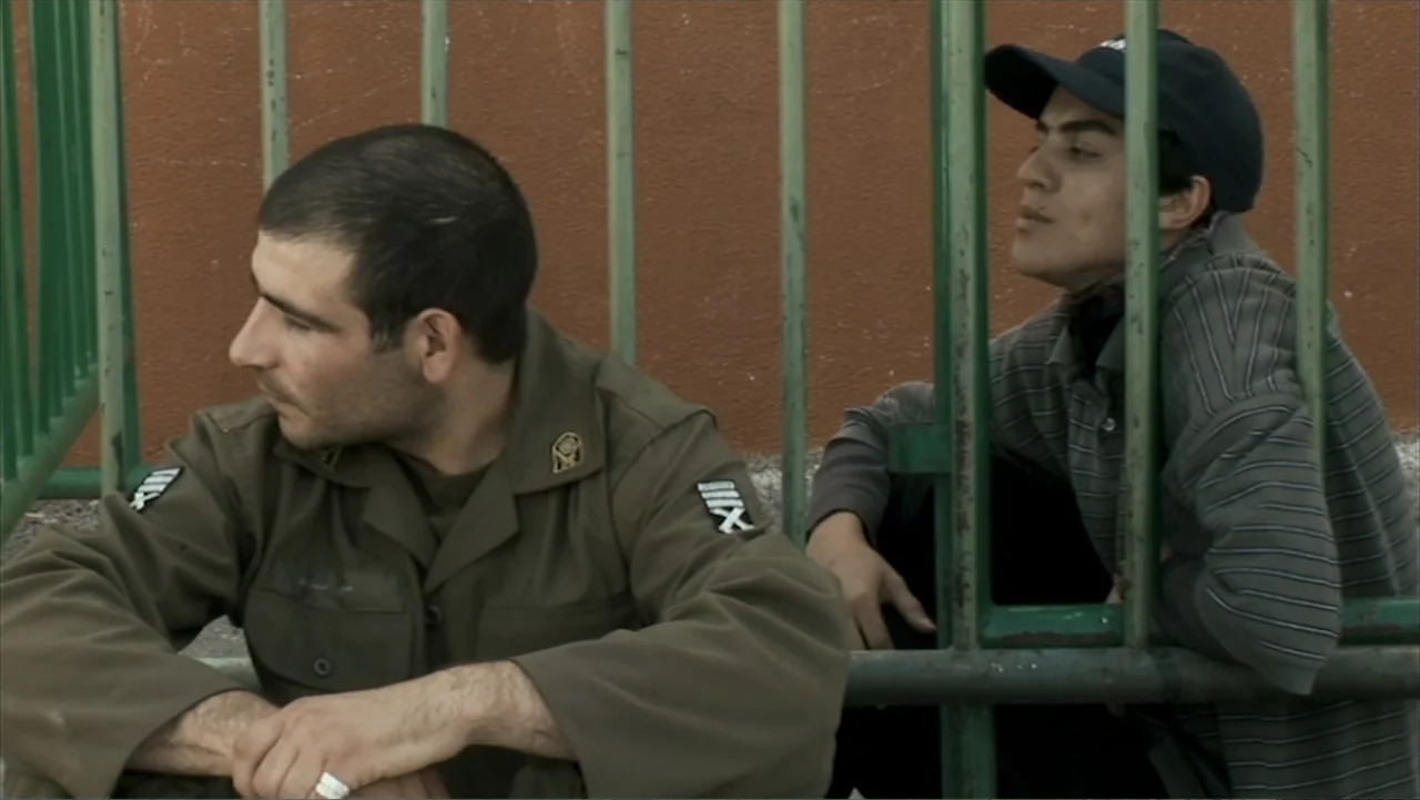 Offside (2015) movie still: medium — An outdoor area with a brown painted wall behind a green metal fence. Two men in casual and military attire sit…