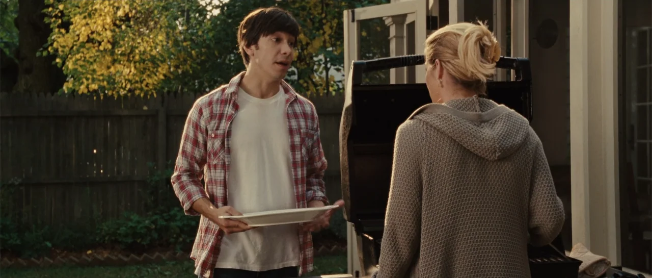 Going the Distance (2010) movie still: medium — A suburban backyard features a wooden privacy fence and golden autumn foliage. A man holding a plate…