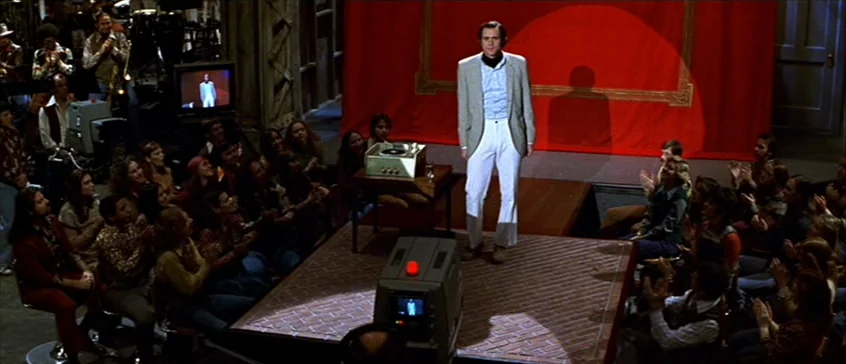 Man On The Moon (1999) movie still: wide — A television studio setting features a small platform with a bright red background. A man stands on the…