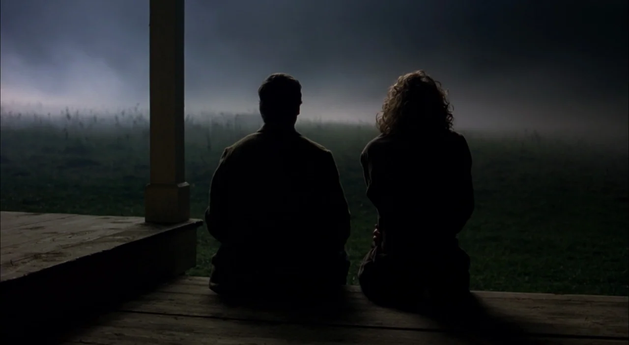 The Village (2004) movie still: medium — A man and a woman sit together on the wooden edge of a porch. They observe a dark, foggy field stretching…