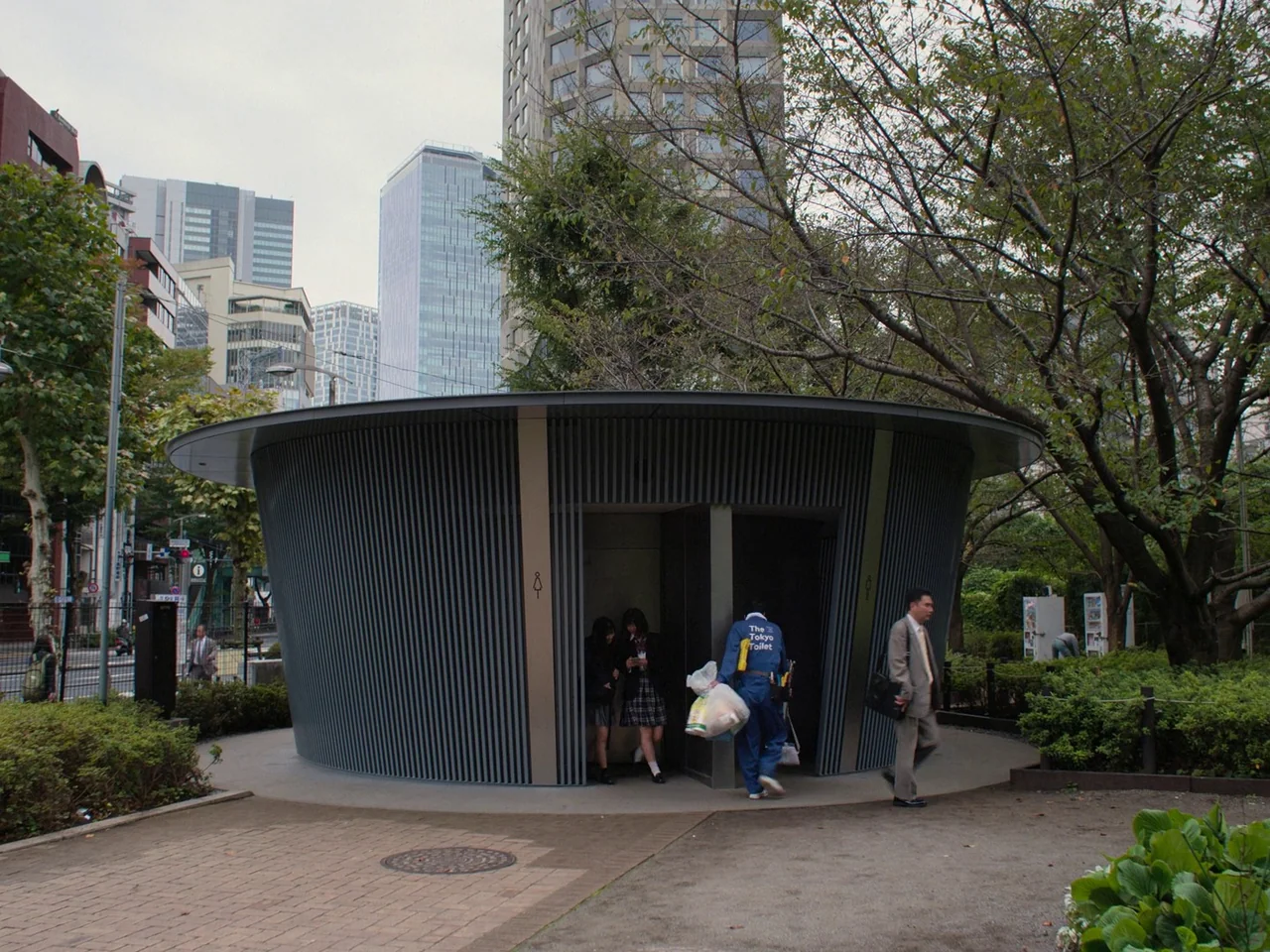 Perfect Days (2023) movie still: wide — A circular modern restroom building sits in a public city park surrounded by trees. People walk near the…