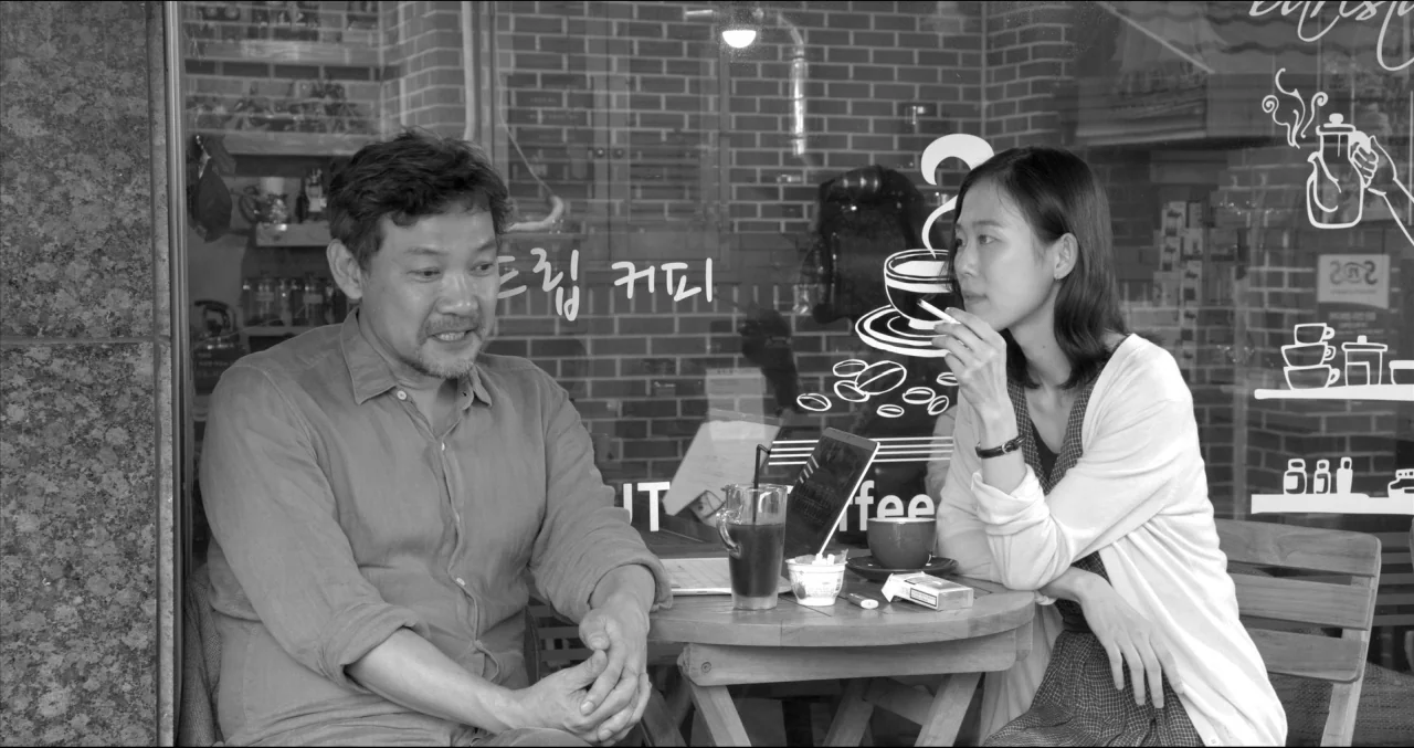 Grass (2018) movie still: medium — A man and a woman sit outside a shop with white graphic decals on glass. The man faces the woman at a small round…