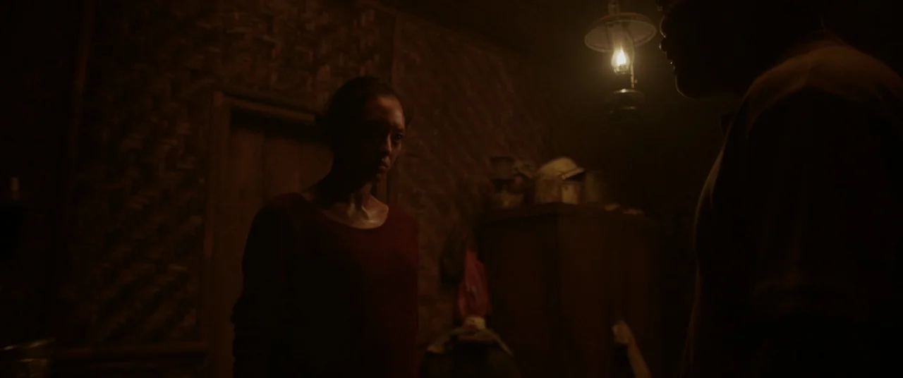 Impetigore (2019) movie still: medium — A dimly lit room with woven bamboo walls is illuminated by a hanging lamp. A woman in a red shirt stands…