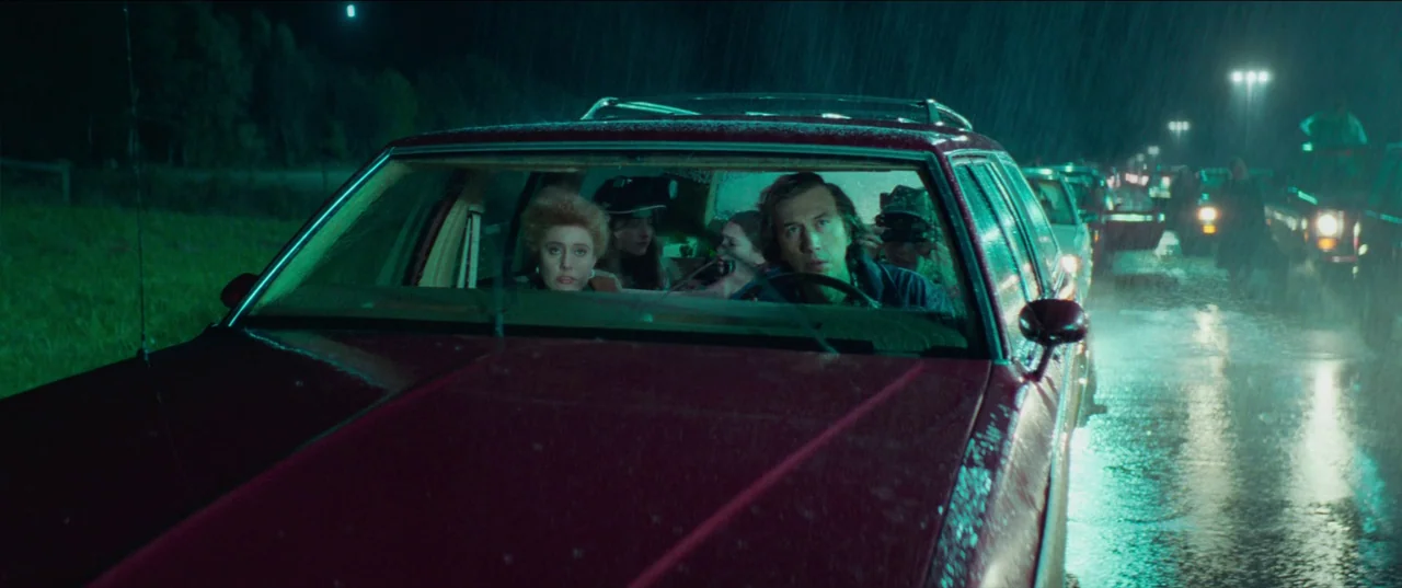 White Noise (2022) movie still: medium — A dark, rain-slicked highway at night with a line of stopped vehicles under streetlights. A maroon sedan…