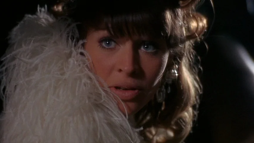 Petulia (1968) movie still: closeup — A dark environment with a woman wearing a white feather boa around her neck. She looks off-screen with wide…