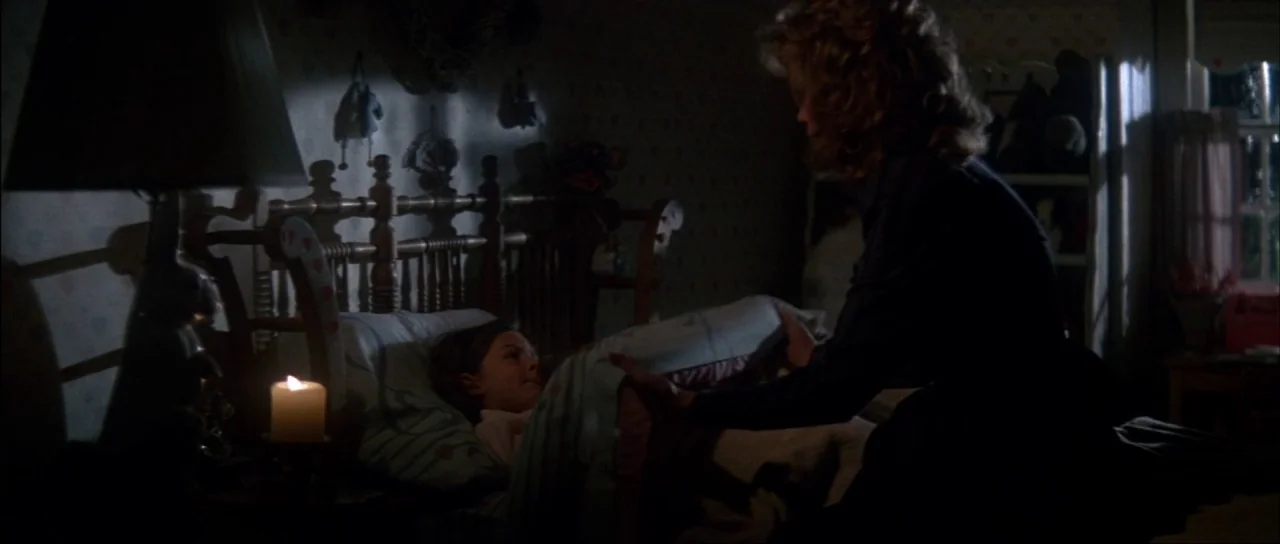 The Monster Squad (1987) movie still: medium — A dimly lit room features a wooden bed frame against a wallpapered wall. A woman leans over to pull a…
