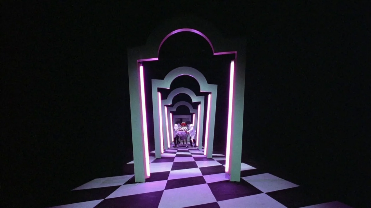 Pee-Wee’s Big Adventure (1985) movie still: wide — A dark hallway features a series of repeating white arches illuminated by pink neon lights. Three…