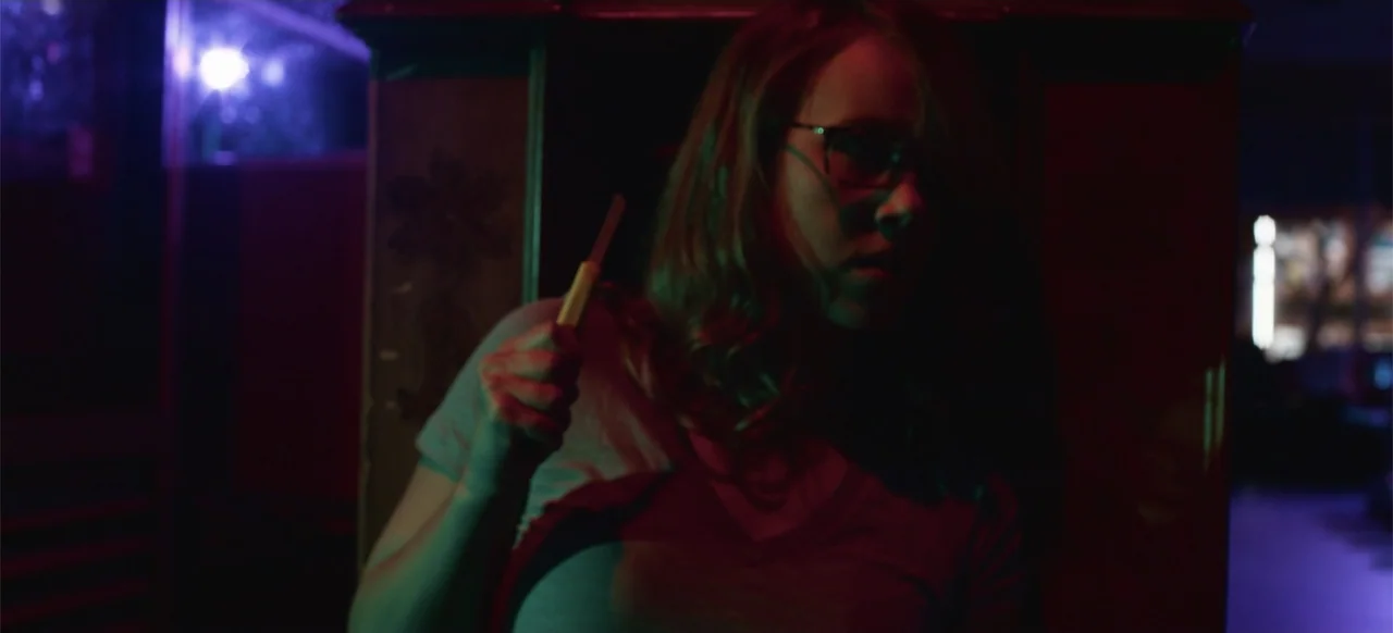 Hellbreeder (2004) movie still: medium — A dimly lit room at night illuminated by harsh blue and red neon lights. A young woman with glasses holds a…