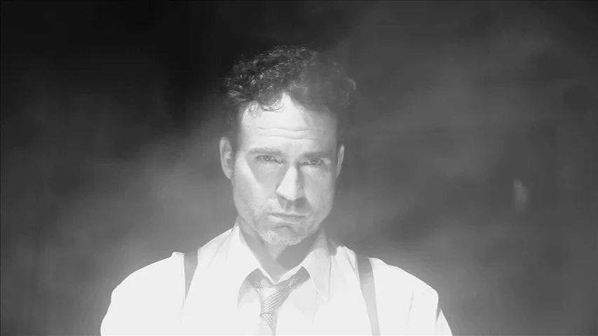 Keyhole (2011) movie still: closeup — A man stands in a dimly lit, undefined interior space filled with light haze. He wears a dress shirt and…