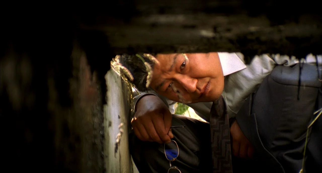Memories of Murder (2003) movie still: closeup — A man peers into a narrow hole between concrete edges in a grassy area. He holds a pair of glasses…