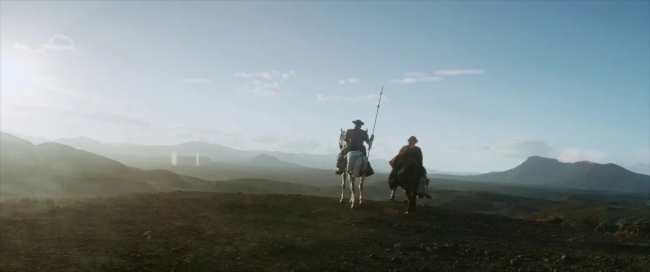 The Man Who Killed Don Quixote (2018) movie still: wide — A vast, arid desert landscape features distant mountains under a pale, hazy blue sky. Two…