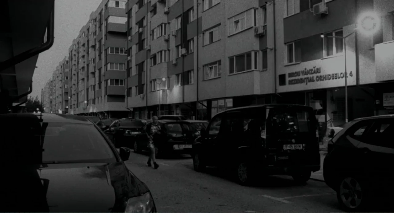 Do Not Expect Too Much from the End of the World (2023) movie still: wide — A dense urban street at night lined with parked cars and apartment…