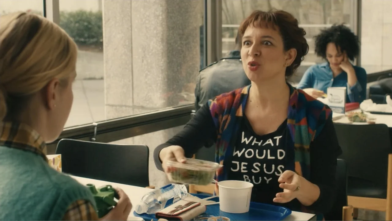 Maggie's Plan (2016) movie still: medium — An interior cafeteria setting with tables arranged in front of large windows. A middle-aged woman sits at…