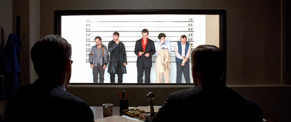 The Usual Suspects movie still: wide — A dimly lit office overlooks a bright, white police lineup room through a glass window. Five men stand in a…