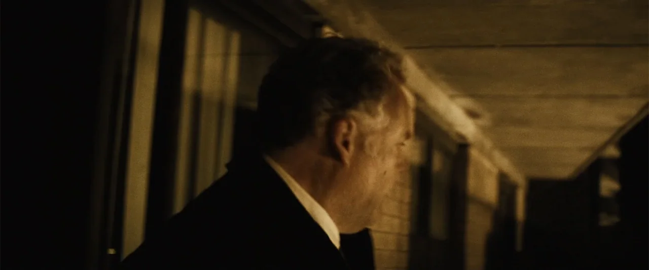 Run All Night (2015) movie still: closeup — A dark outdoor motel corridor at night illuminated by harsh artificial light sources. A middle-aged man…